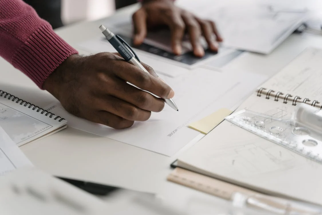 Person sketching and writing notes on architectural plans at a desk with drafting tools
