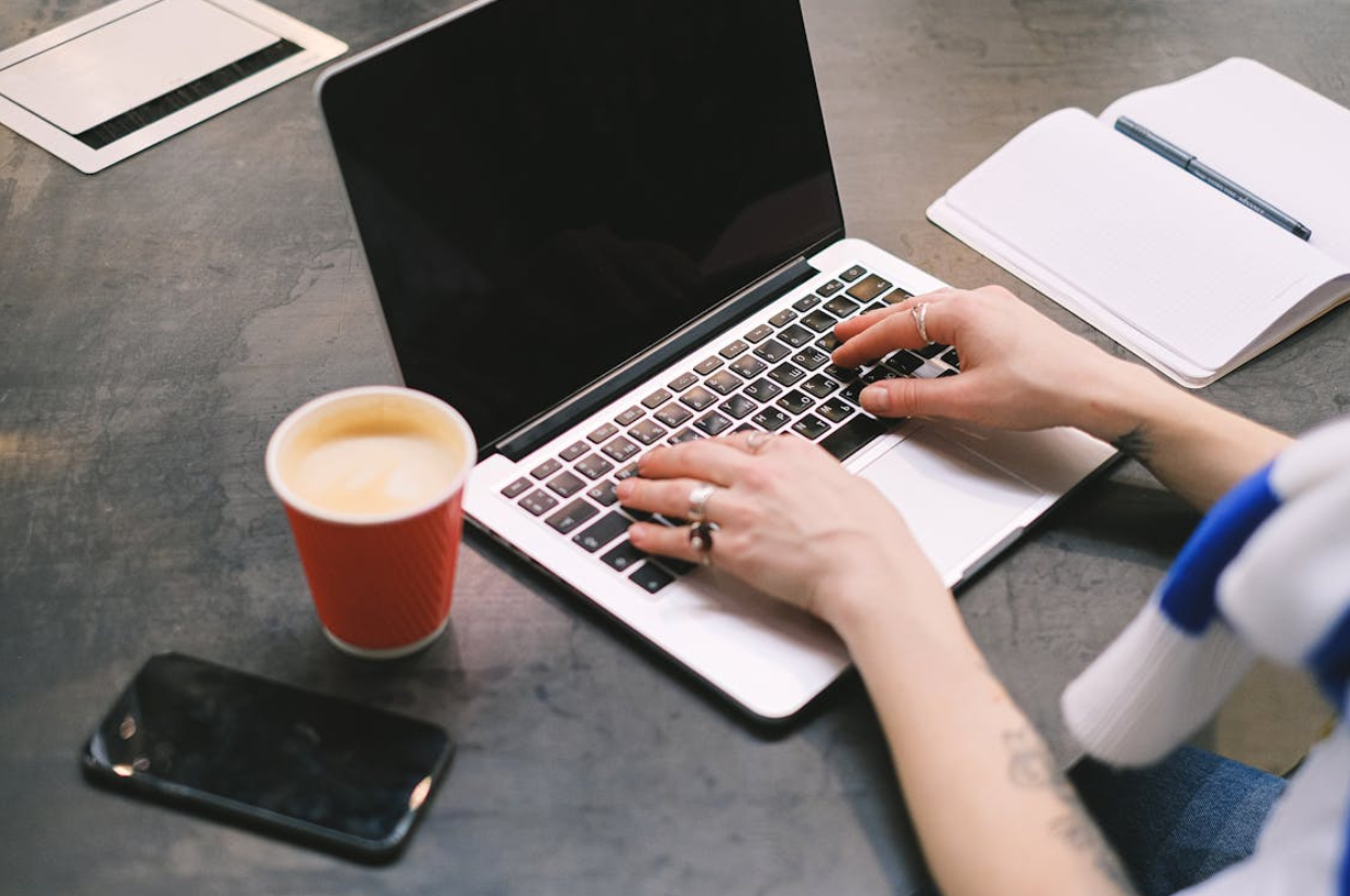 A person typing on a laptop with a notebook, coffee cup, and smartphone on the table