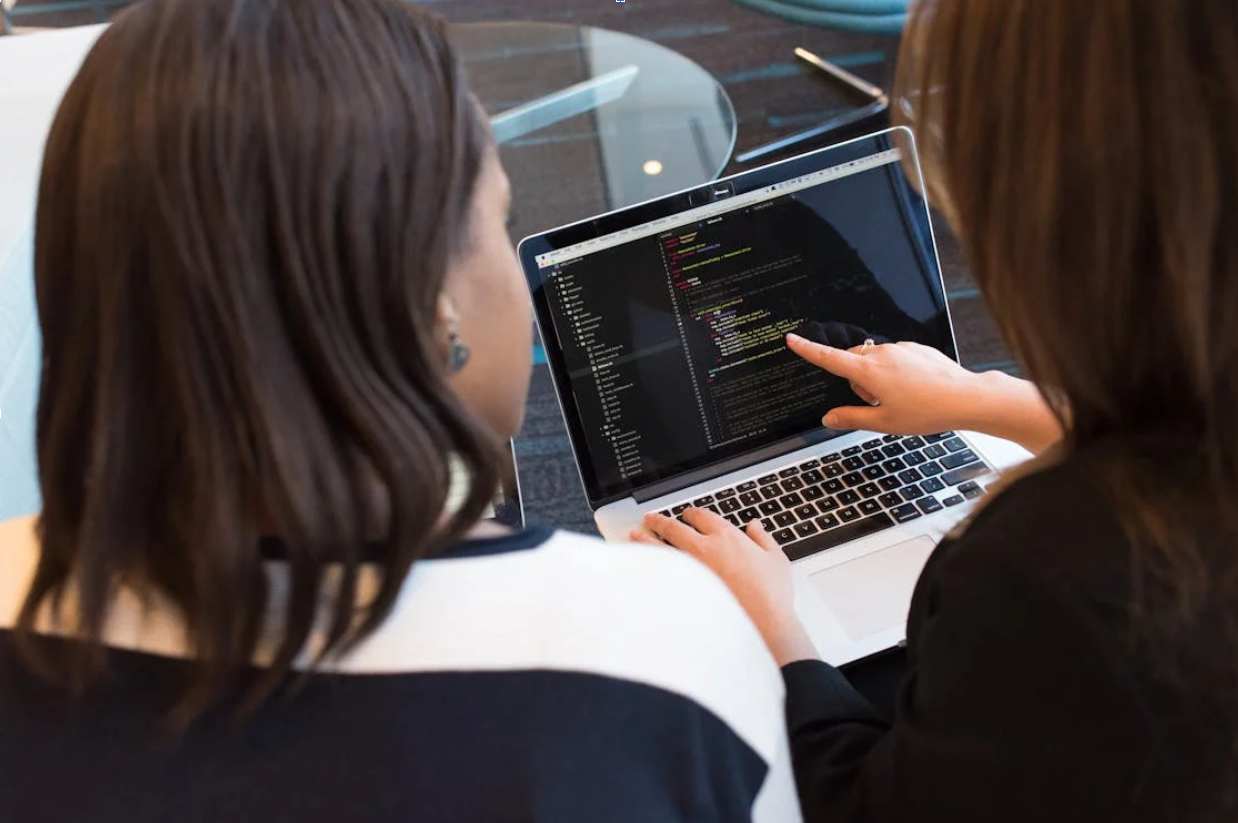 Two women reviewing and discussing code on a laptop in a collaborative workspace