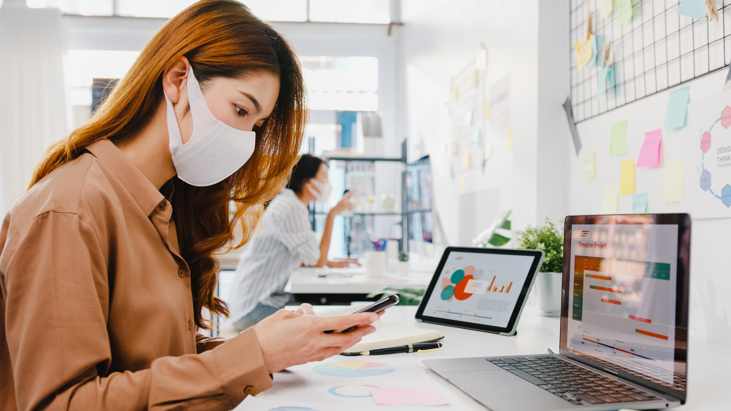 A woman in a face mask working on a phone and laptop in an office