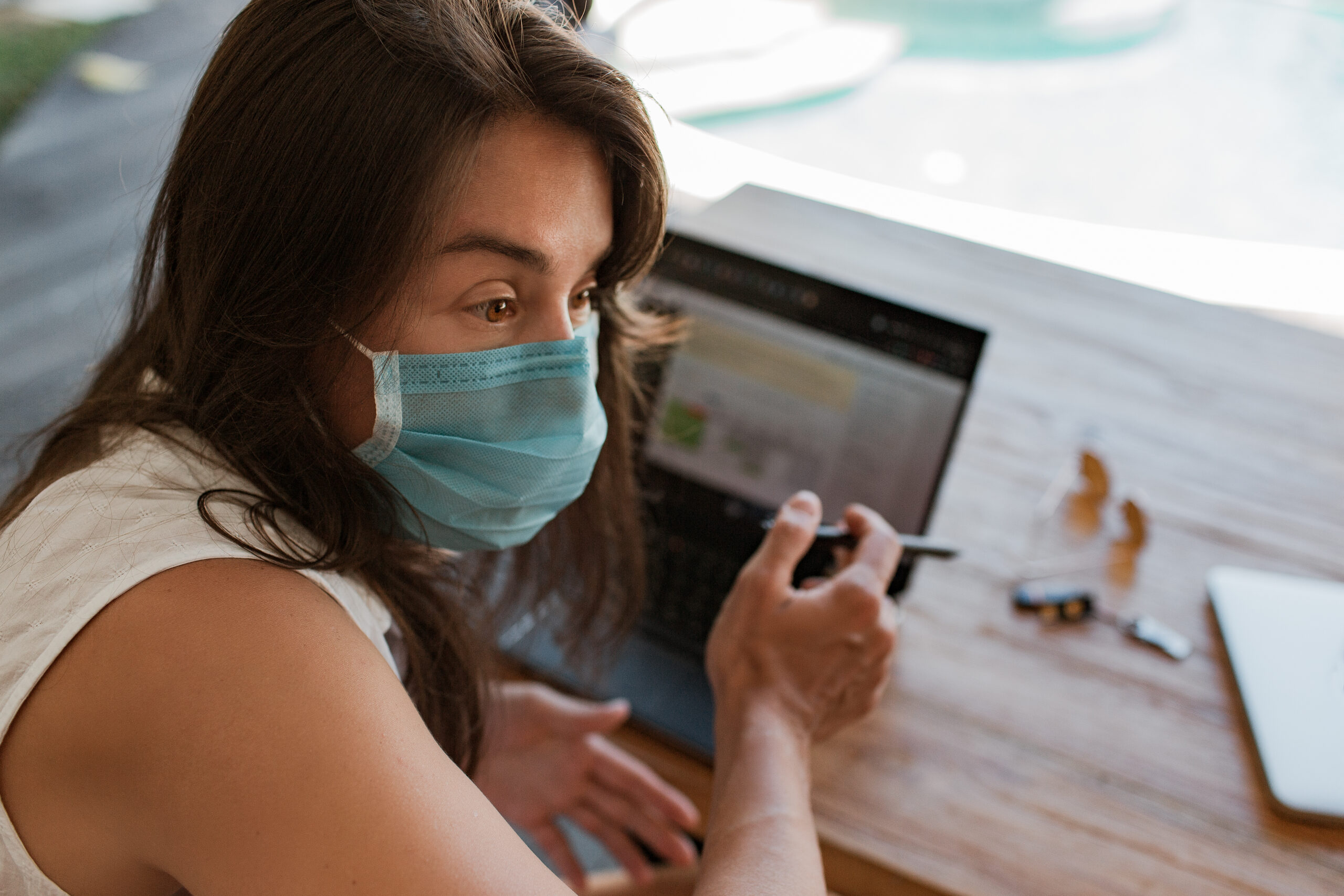 A woman wearing a face mask working on a laptop and holding a pen