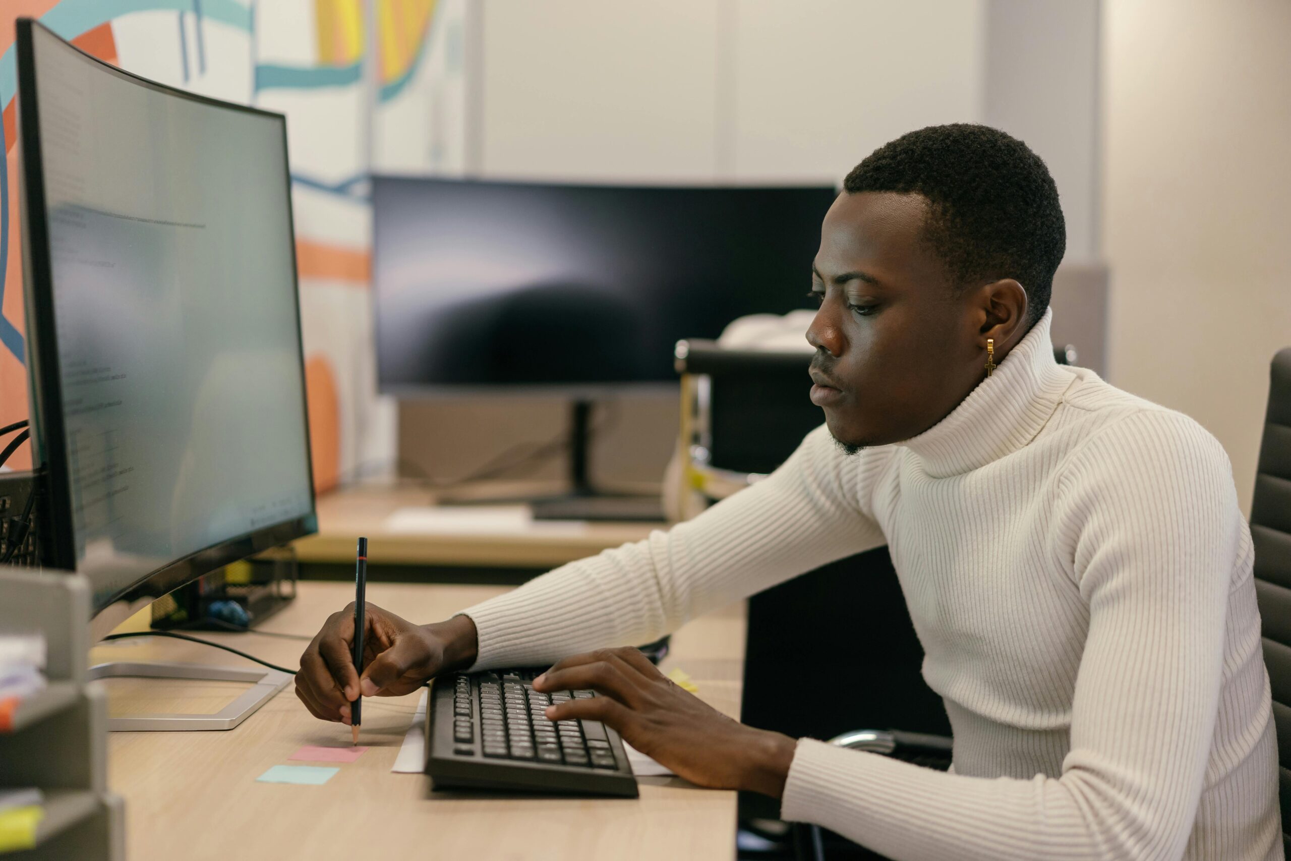 A man working at a desk with a large monitor, taking notes while using a keyboard